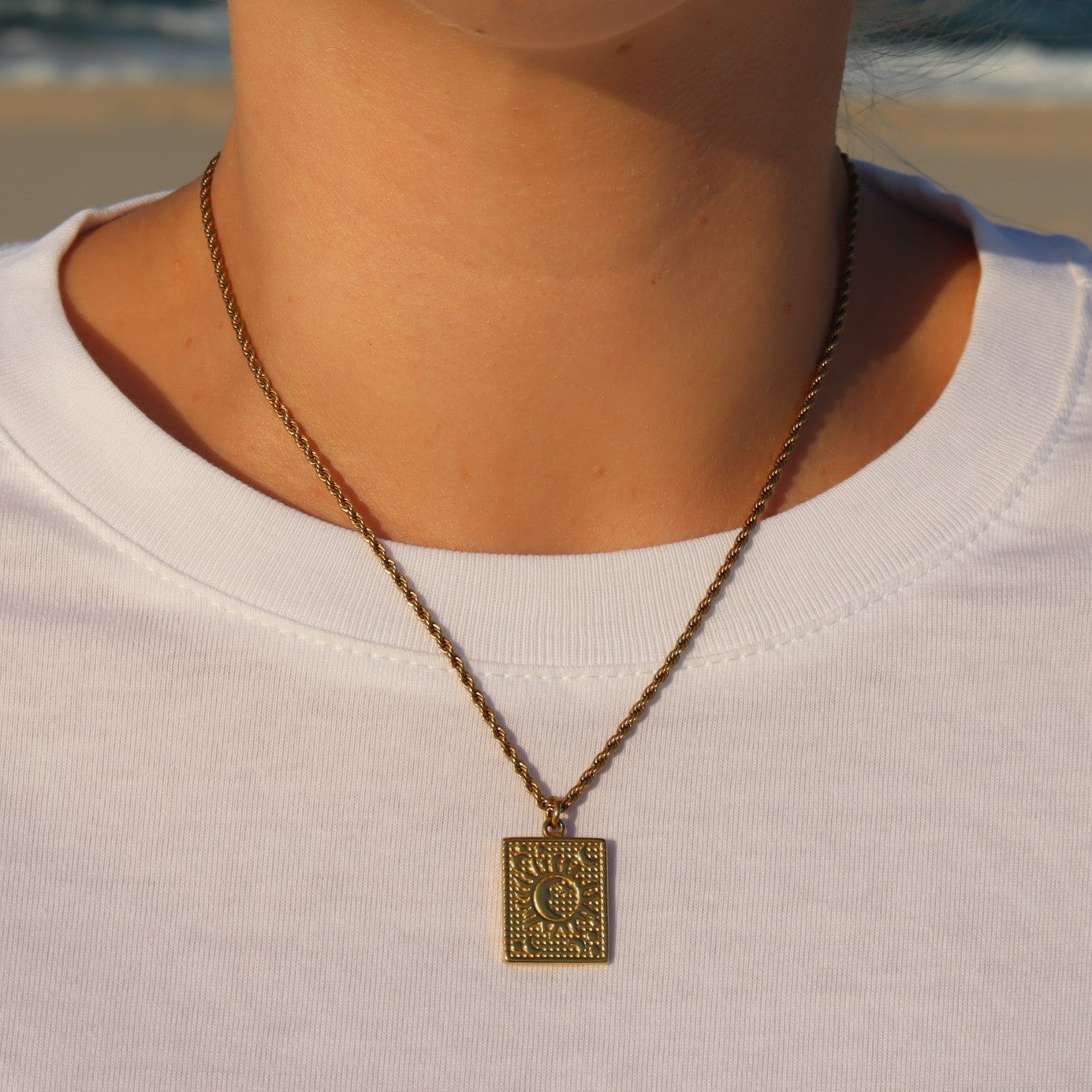 Gold necklace with a rectangular pendant worn by a person on a beach.
