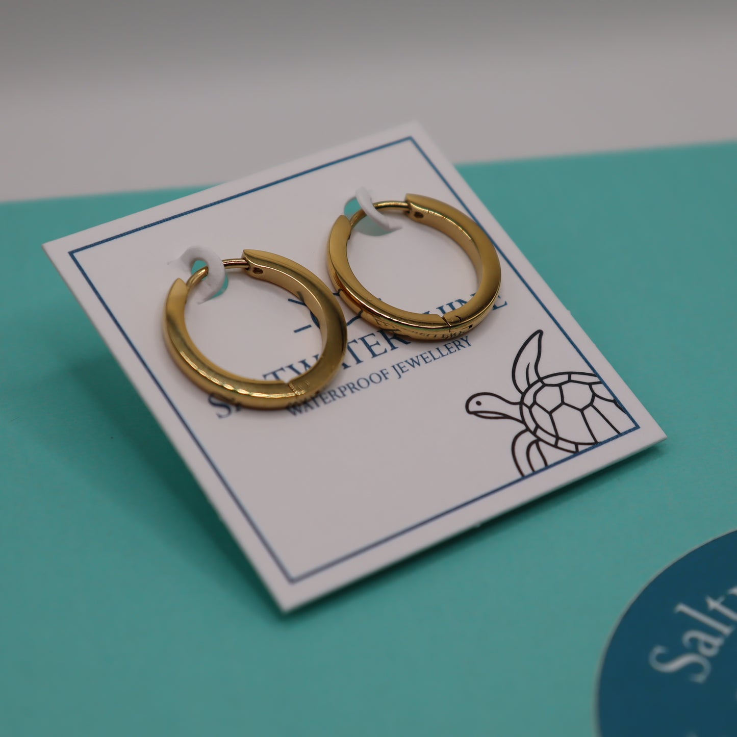 Gold hoop earrings on a branded card with a turtle design against a teal background.