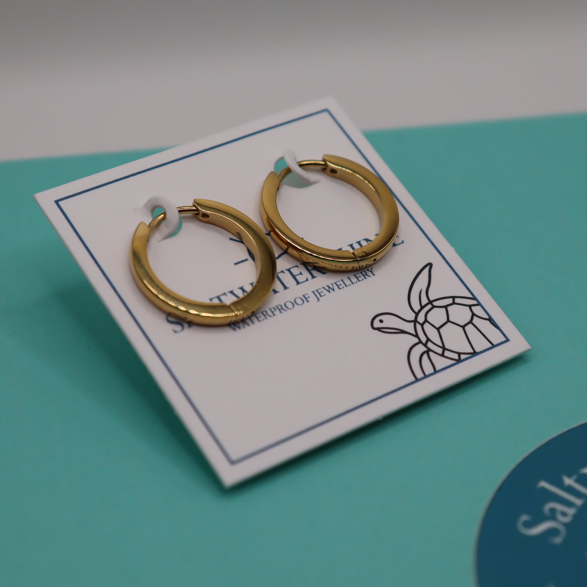 Gold hoop earrings on a branded card with a turtle design against a teal background.