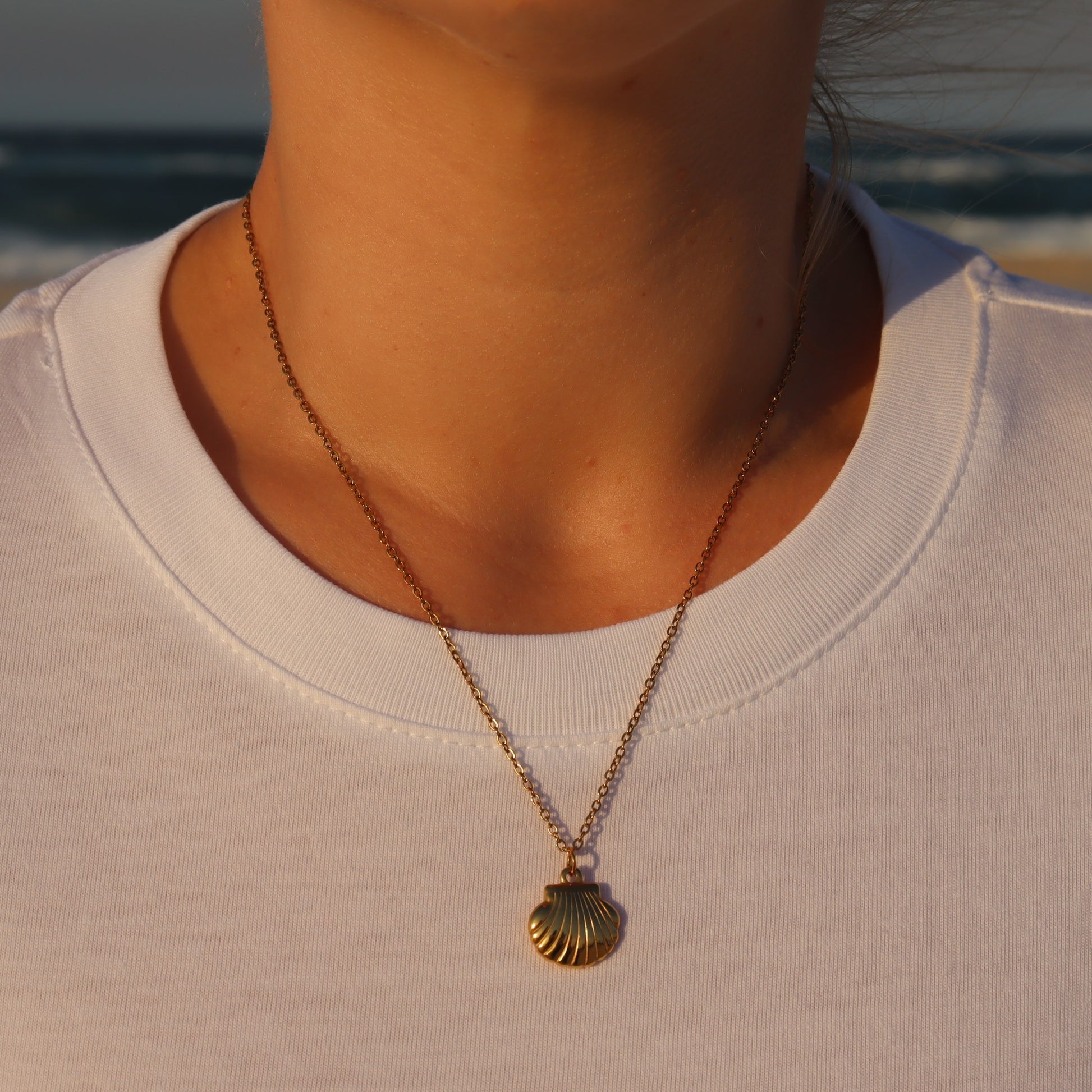 Gold necklace with a shell pendant worn by a person against a blurred natural background