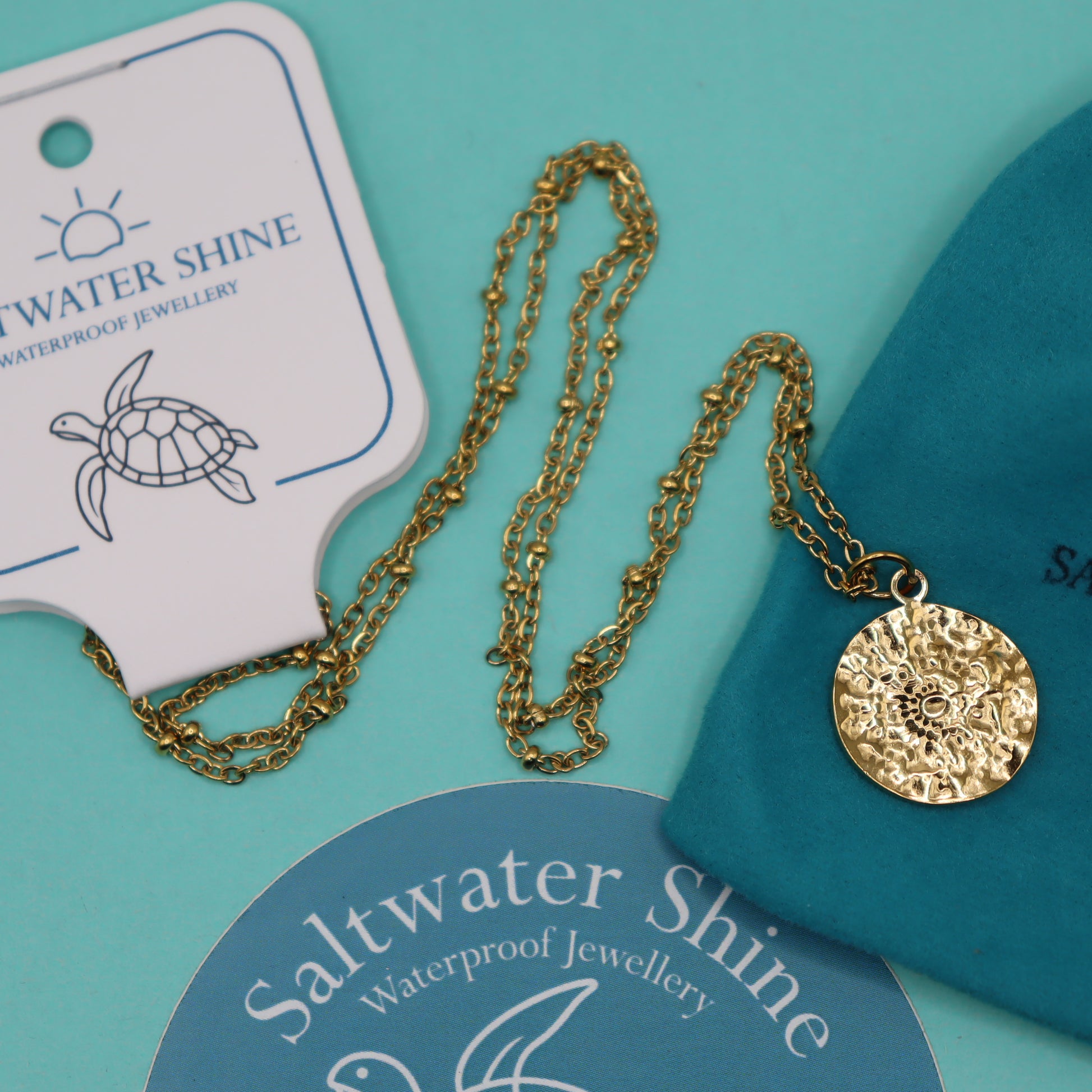 Gold necklace with a textured pendant on a teal background with 'Saltwater Shine' branding.