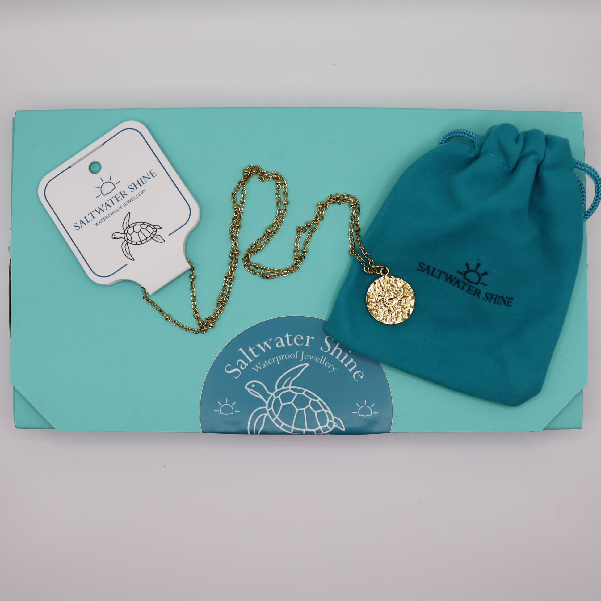 Gold necklace with pendant on a white card, accompanied by a drawstring bag and 'Saltwater Shine' branding.