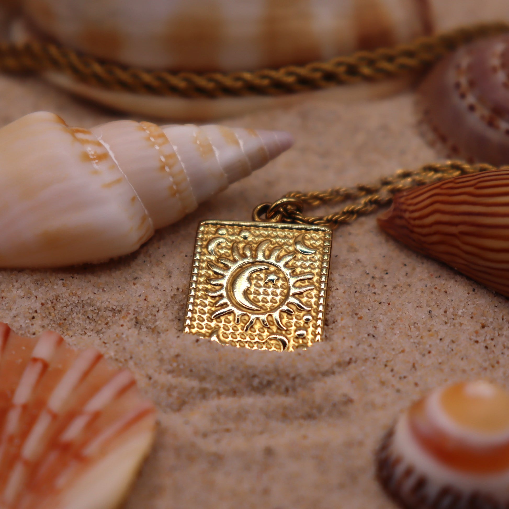 Gold square pendant with sun and moon design on a sandy surface with shells around