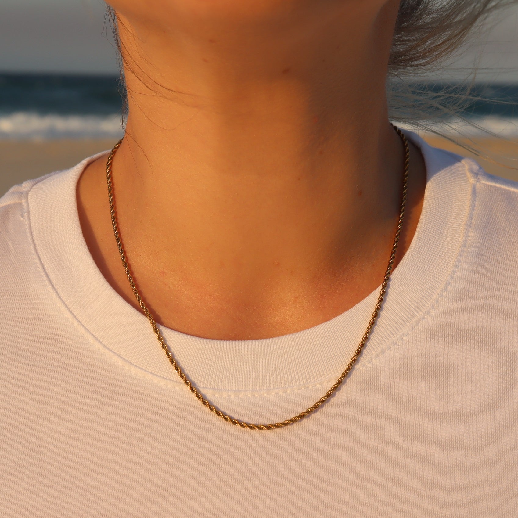 Person wearing a gold rope necklace with a beach in the background at sunset