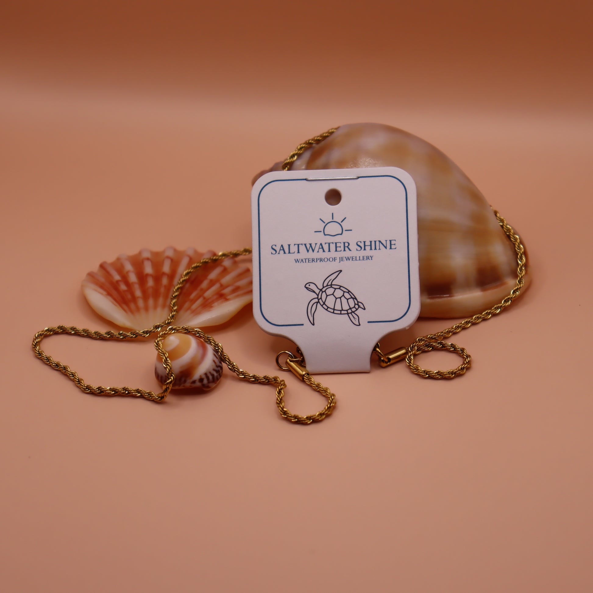 Gold rope necklace with a 'Saltwater Shine' tag and seashells on a peach background