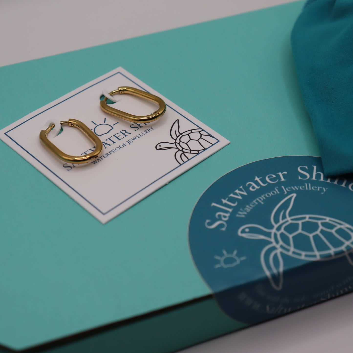 Gold earrings on a card with 'Saltwater Shine' branding on a teal background