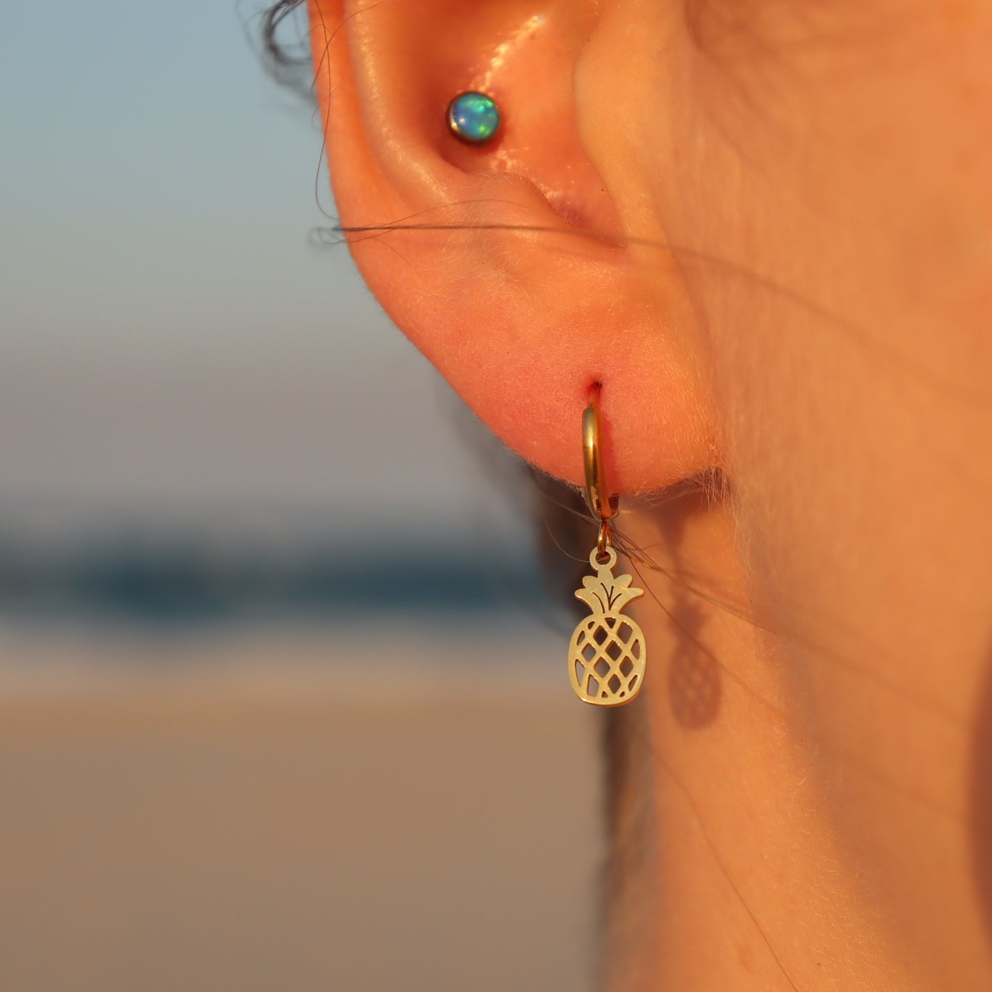 Gold pineapple earring worn in an ear with a blurred beach background