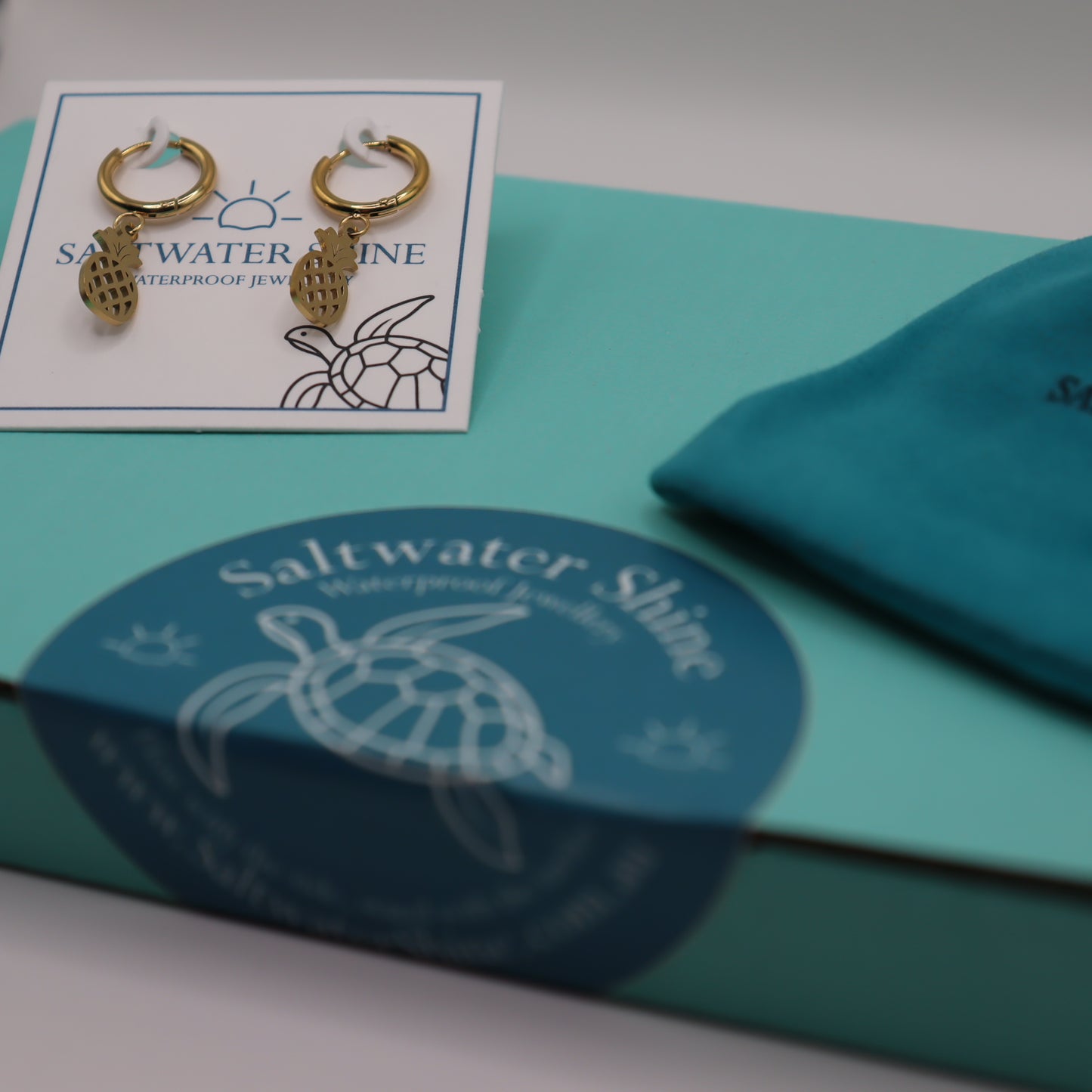 Gold pineapple earrings on a 'Saltwater Shine' branded card with packaging in the background.