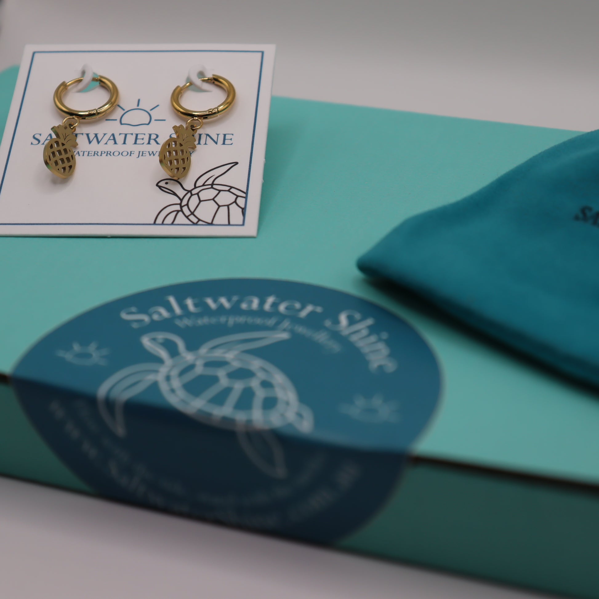 Gold pineapple earrings on a 'Saltwater Shine' branded card with packaging in the background.