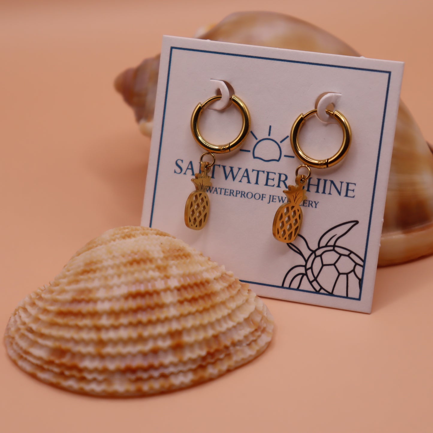 Gold pineapple earrings with a turtle logo on a 'Saltwater Shine' card, surrounded by seashells.