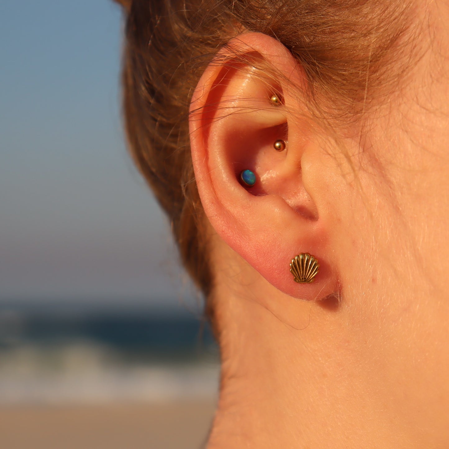 Close-up of an ear with multiple earrings, one of the shell stud, set against a blurred beach background