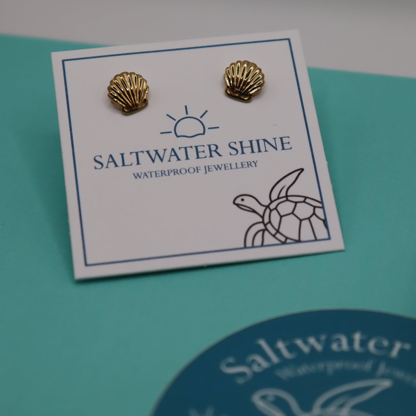 Gold shell-shaped earrings on a 'Saltwater Shine' branded card against a teal background.