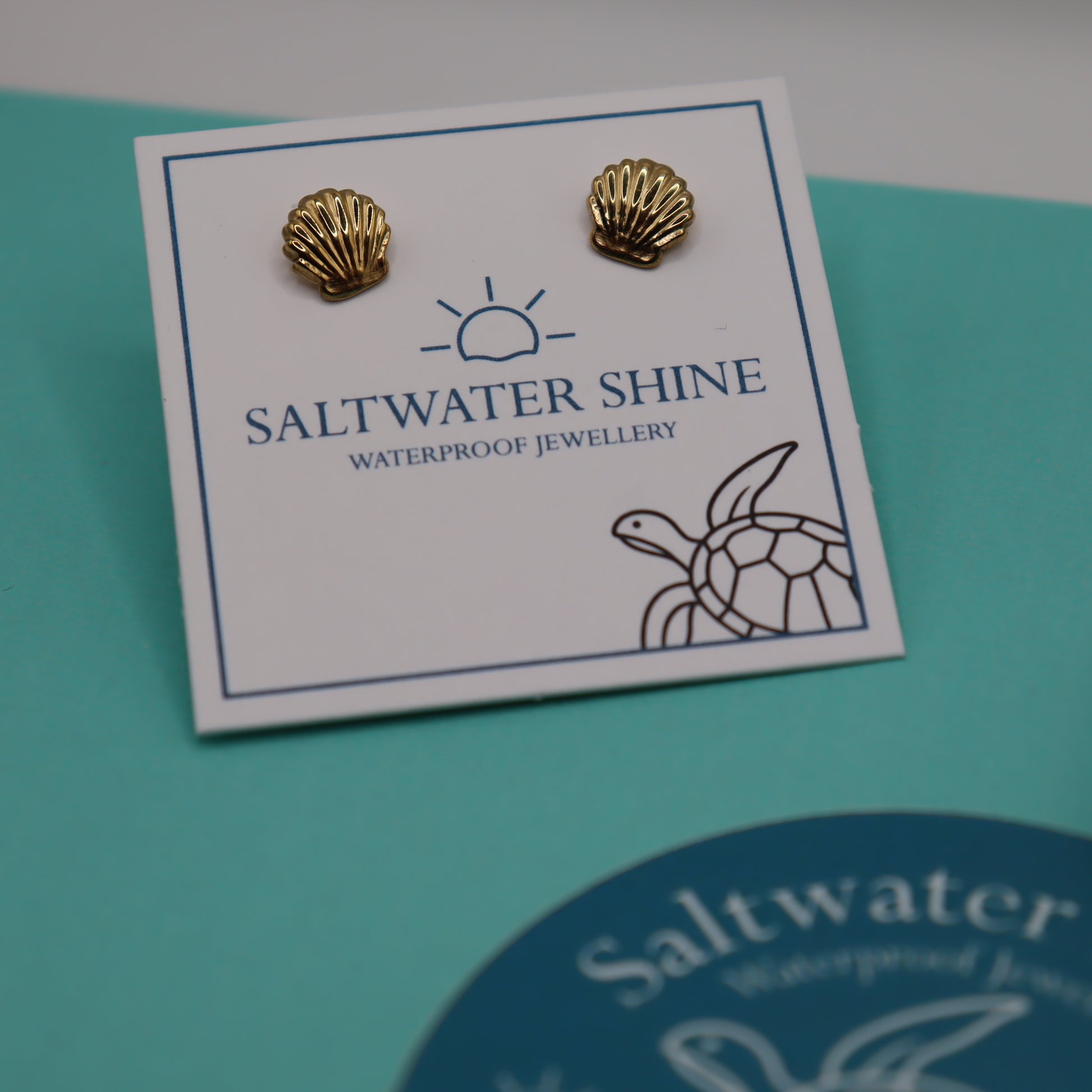 Gold shell-shaped earrings on a 'Saltwater Shine' branded card against a teal background.