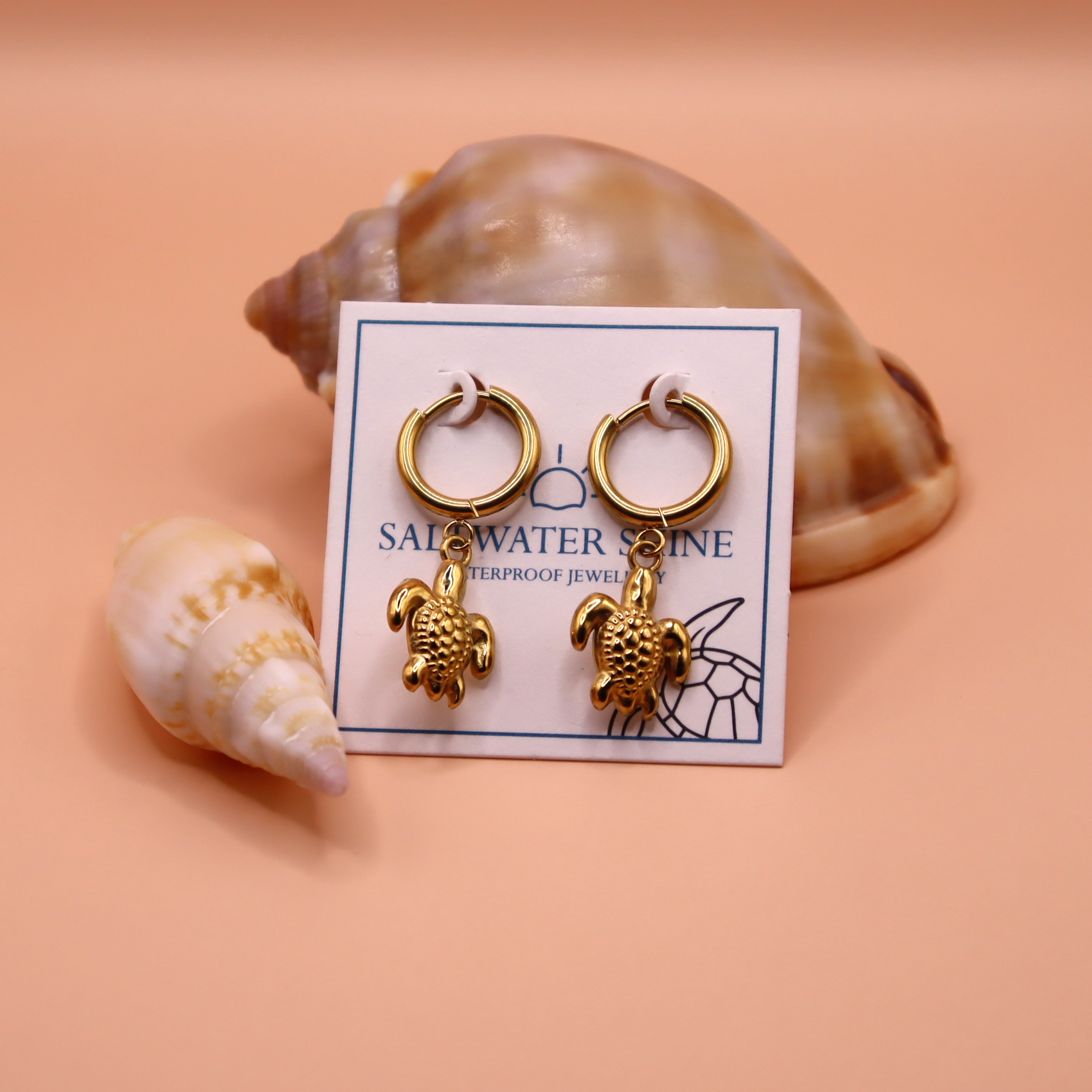 Gold turtle earrings on a 'Salwater Shine' card with seashells in the background.