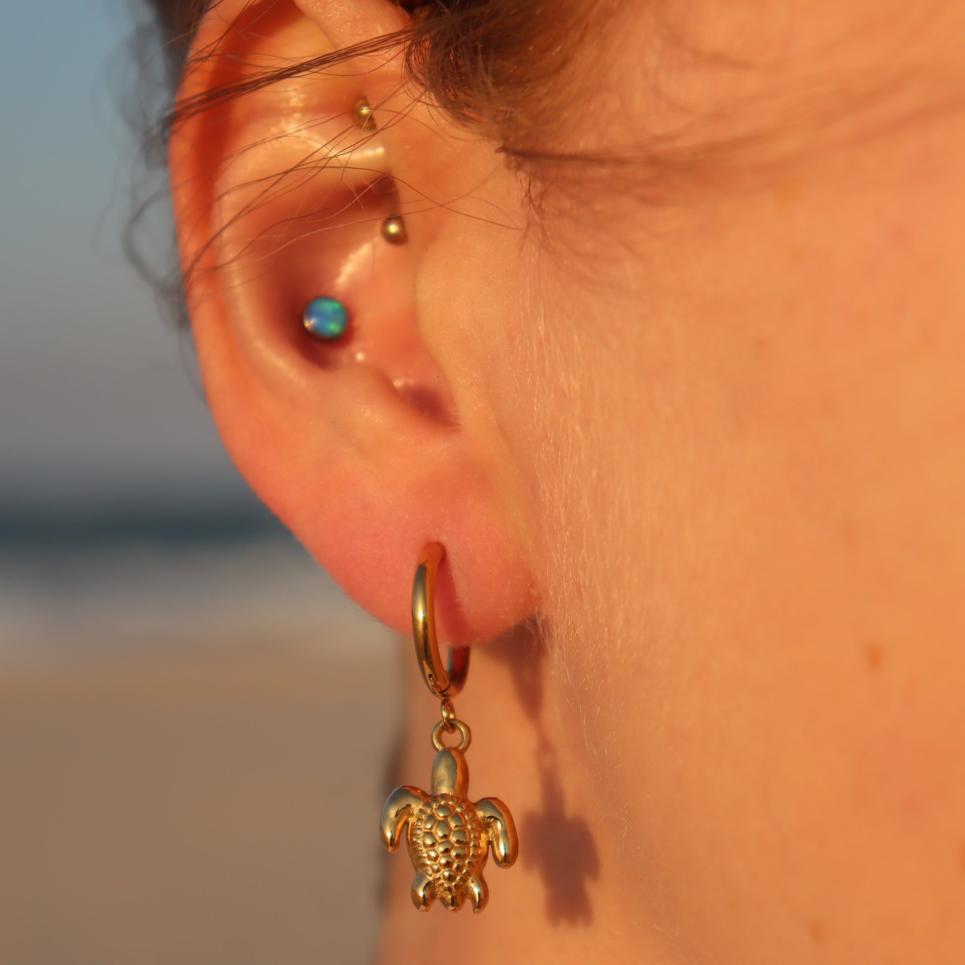 Close-up of an ear with a gold turtle earring against a blurred beach background