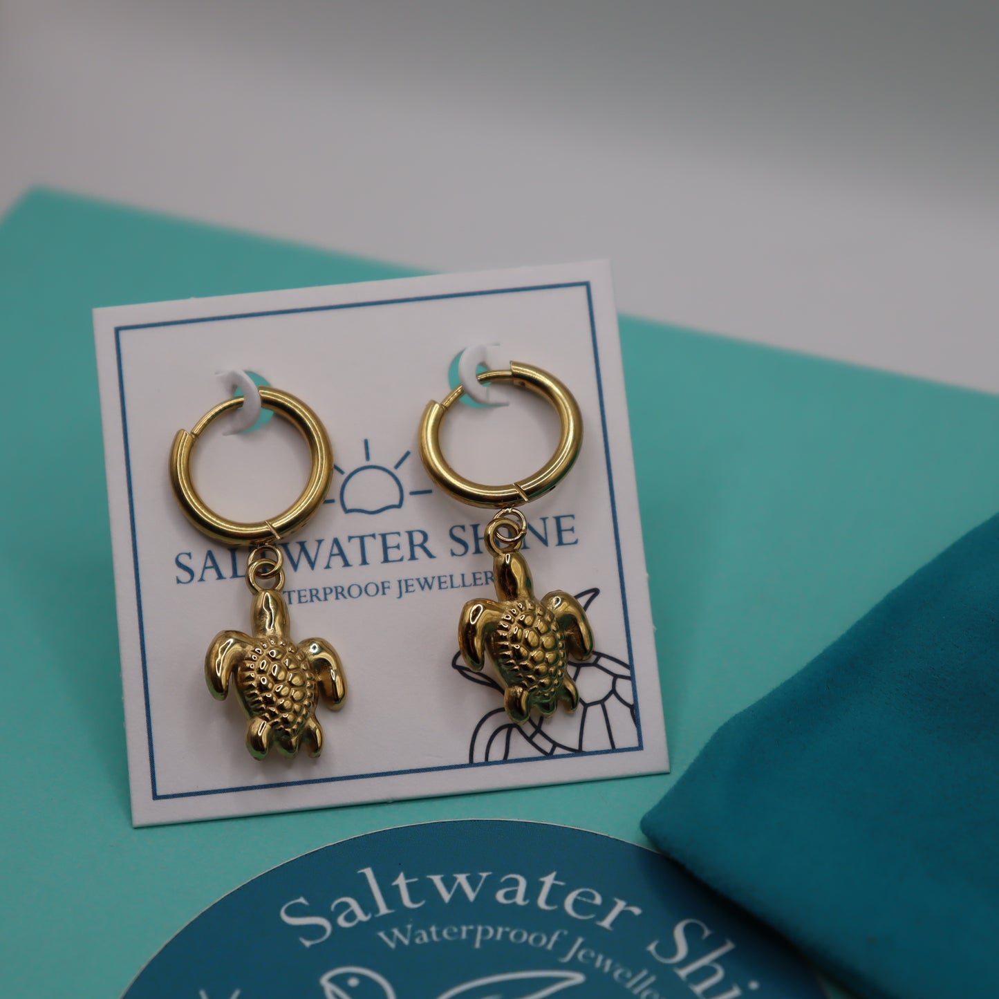Gold turtle earrings on a 'Saltwater Shine' branded card against a teal background
