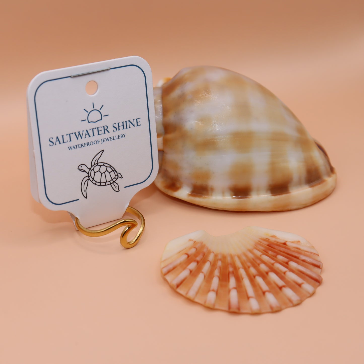 Gold ring with 'Saltwater Shine' tag and two seashells on a peach background